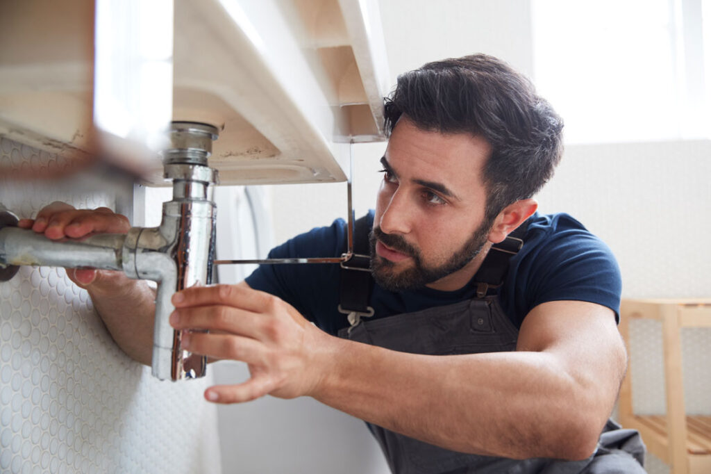 A male plumber in overalls fixing a leaking pipe under a sink in a home bathroom.